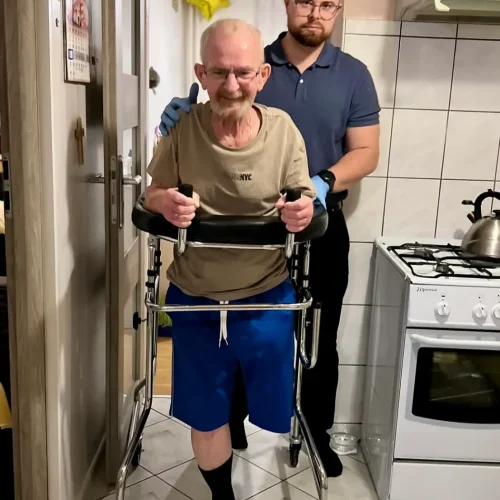 Starszy mężczyzna po amputacji nogi poruszający się z balkonikiem po mieszkaniu, w towarzystwie fizjoterapeuty.
