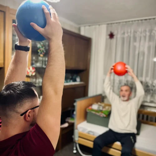 Mężczyzna w trakcie rehabilitacji, siedzący na łóżku z uniesionymi nad głowę rękami w których trzyma czerwoną piłkę.