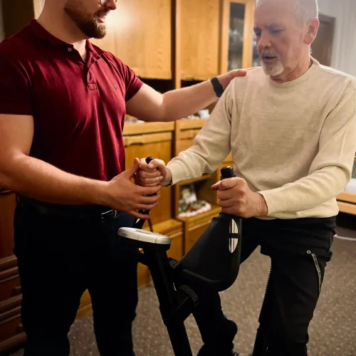 Starszy mężczyzna w pokoju na rowerku rehabilitacyjnym w towarzystwie fizjoterapeuty.